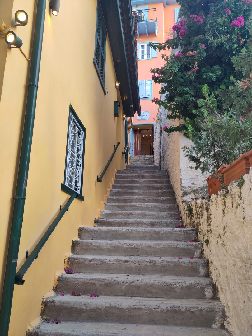 Steps leading up a narrow alley in Nafplio, flanked by a pastel yellow wall on the left and flowering green plants on the right.