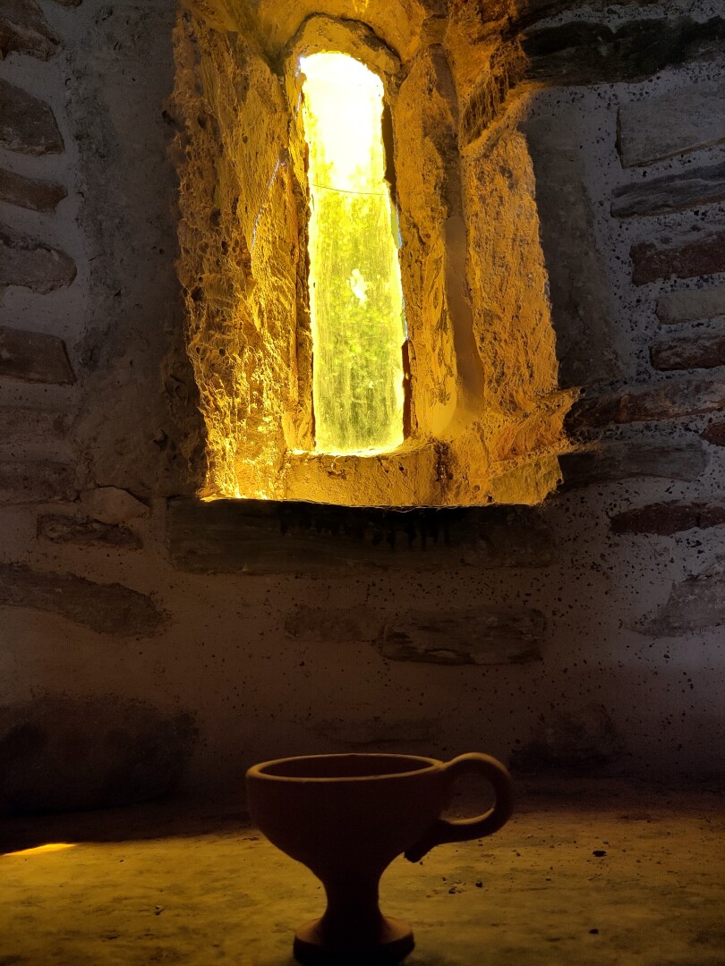 A more artistic upwards angle of a tiny ceramic cup with a single handle, sitting on a stone slab silhouetted in the yellowish light pouring in from the small tall and narrow window behind it.