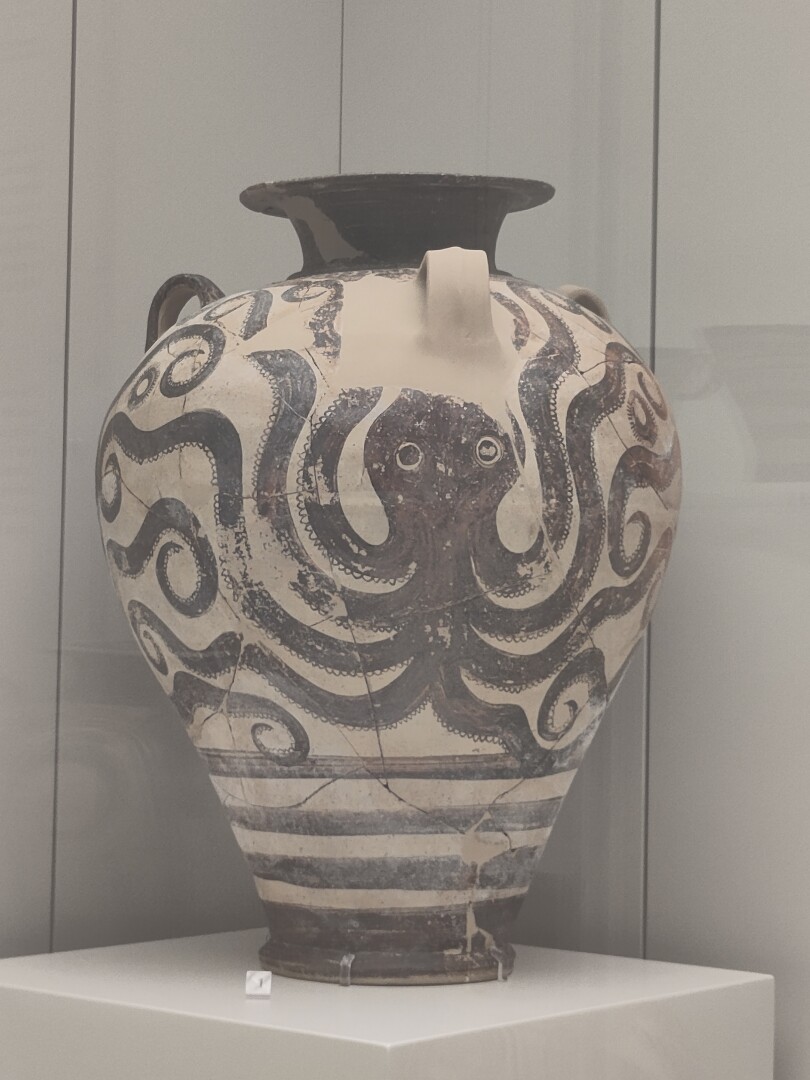 A tall white vase with three tiny handles around the top and a big ol' octopus figure drawn on it in black, four tentacles on each side curving upwards to flank its head.