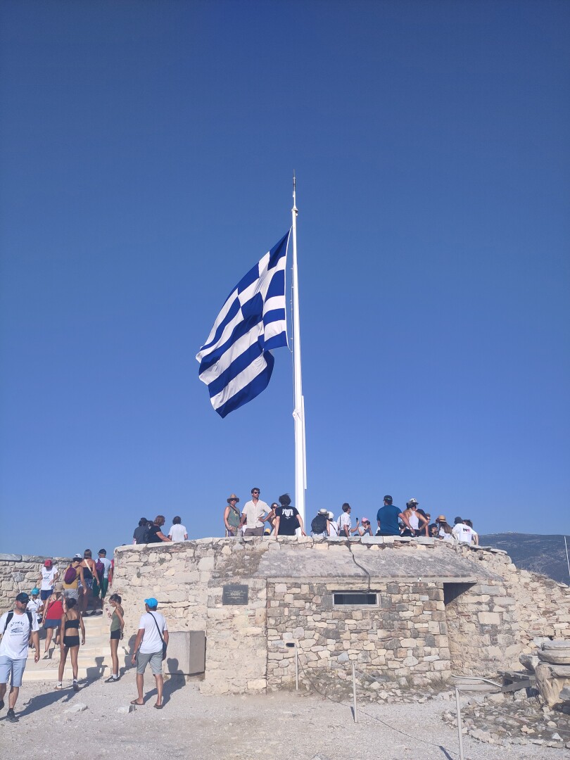 The Greek flag flying against clear blue skies on the Athenian Acropolis, with lots of people around it admiring the view of the surrounding city.