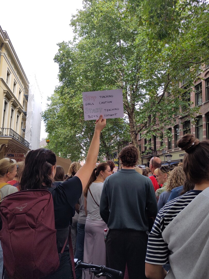Several people standing in the street with their backs towards the camera, one person holding up a sign that says:
"Stop teaching girls caution. Start teaching boys consent."