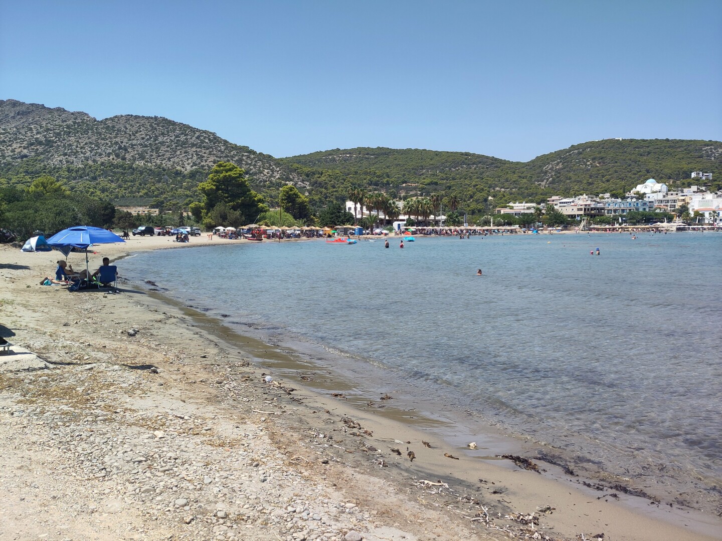 A mostly sandy somewhat rocky beach, bending from left close-up to right background with green hills and some town buildings, hugging the clear blue waters of the Aegean sea.