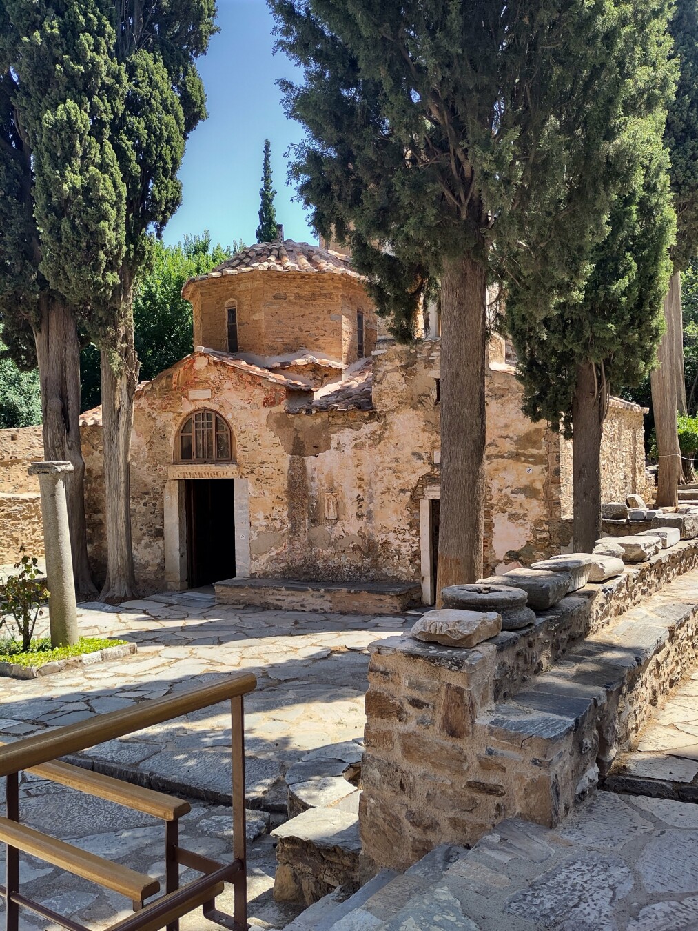 Small byzantine style church in salmon brick with a little dome, flanked by tall cedar trees with a low stone wall and path running along the right side.