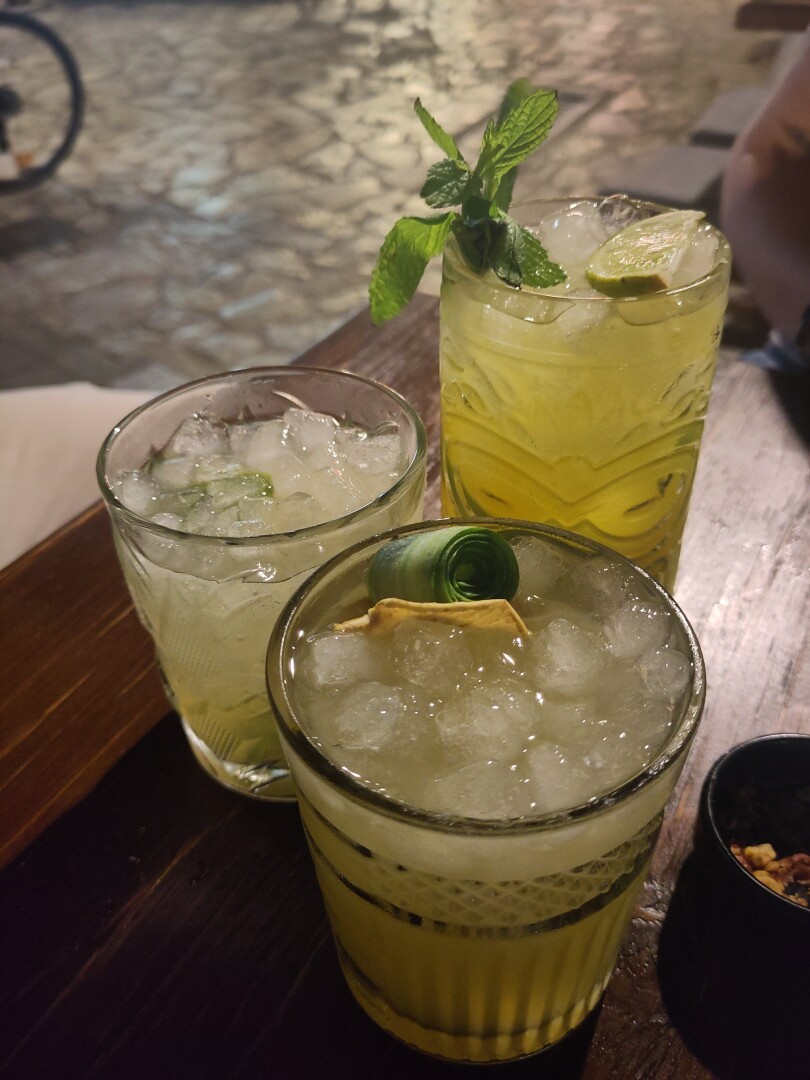 Three more drinks:
On the right is a light green drink in a tall glass with a sprig of mint and slice of lime: passion-fruit no-jito mocktail.
On the left is a tumbler glass with yellow-green drink on crushed ice, a classic caipirinha.
In the middle is another tumbler glass with a different yellow-green drink, this one a Messinian Garden: big sir gin, mastiha liquor, cucumber, lime cordial, apple juice.