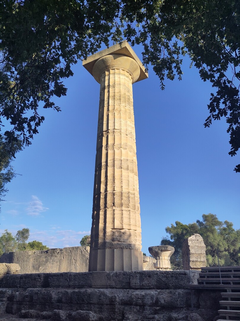 A giant column standing tall against the bright blue sky, framed around the top by an arch of tree leaves, steps leading up on the bottom right. This is the main feature remaining of the Temple of Zeus.