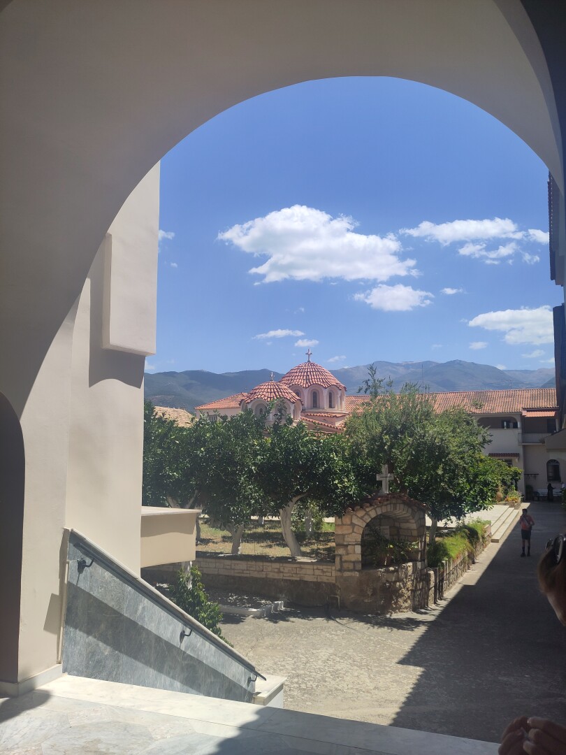 The arched entrance to a monastery forming a natural frame for the inner courtyard, showing a little shrine, some trees in a neat grid and a small domed church in the background.