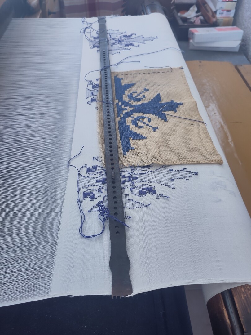 A close-up of another weaving machine, where not only the white silken cloth is woven together, but there is also a still incomplete pattern laid over it in dark blue embroidery. A paper containing the overall pattern sits over the paper as a reference for the maker.