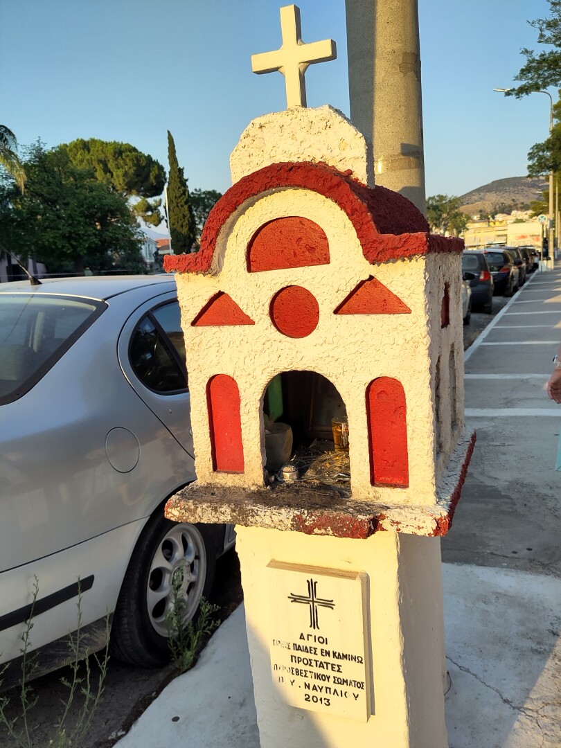 One of the many tiny roadside shrines to be found all across Greece. This one is shaped like a miniature church in white with red windows and roof lining, a small white dome with a cross on top.
