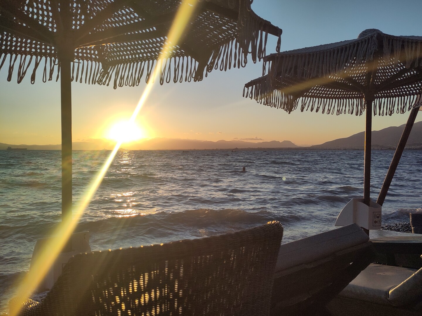 The view of the sun setting on the horizon behind mountains as seen from the beach underneath some parasols, the sun giving off a strong lens flare.