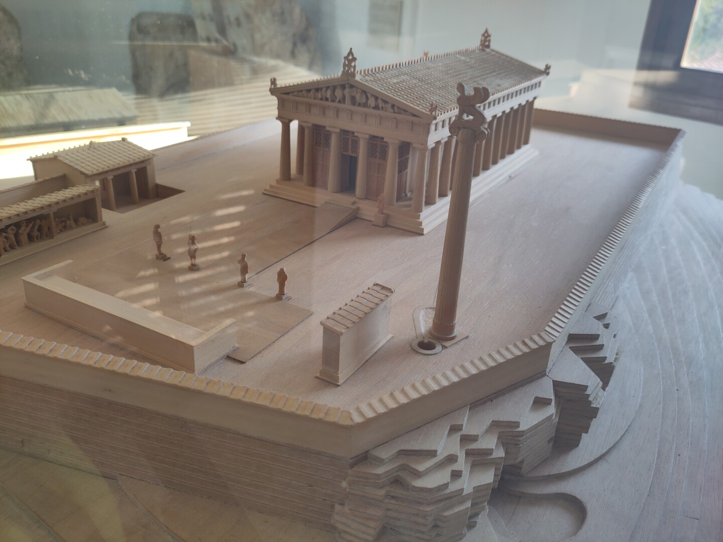 A small model of the Temple of Aphaia, showing how the columned building used to look in the back, including a big statue atop a tall pillar in front and a couple smaller buildings to the side.