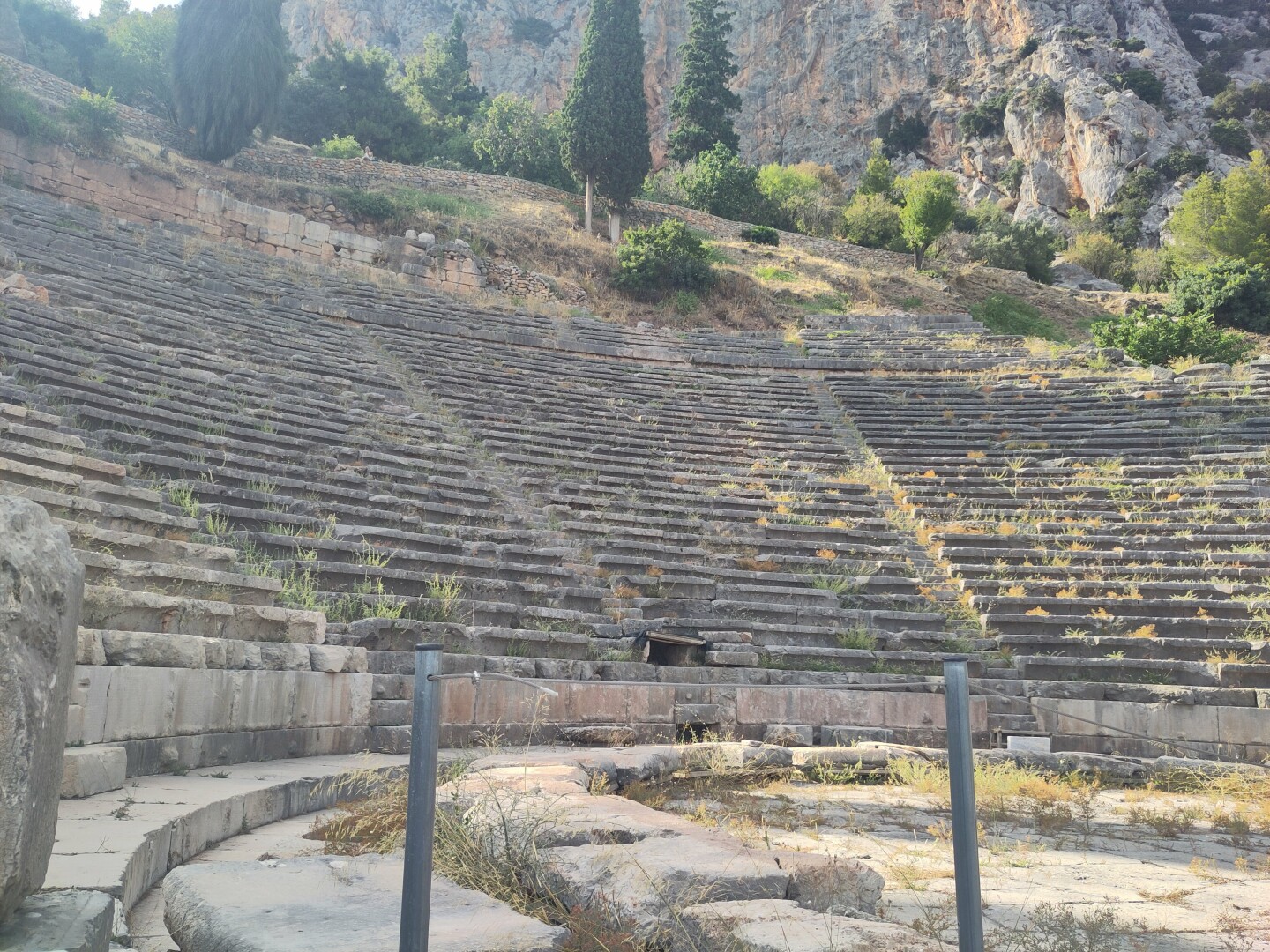 A view of the Ancient Theater in Delphi taken from below to show the half circle stage in front and rows upon rows of stone benches rising up all around it, hugging the mountain side.