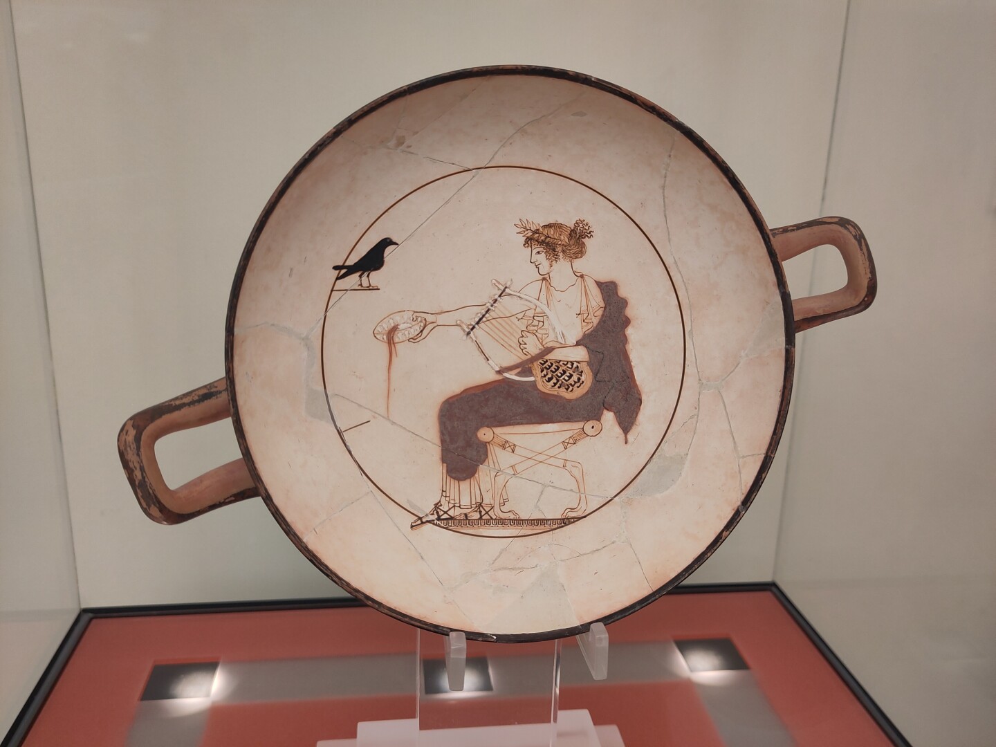 A flat ceramic bowl with a white background and Apollo depicted with a wreath of myrtle leaves, seated on a stool (with lion paws) dressed in white with a red drape over one shoulder. He's holding a lyre with one hand and pouring out wine with the other. A black crow sits in the top left.