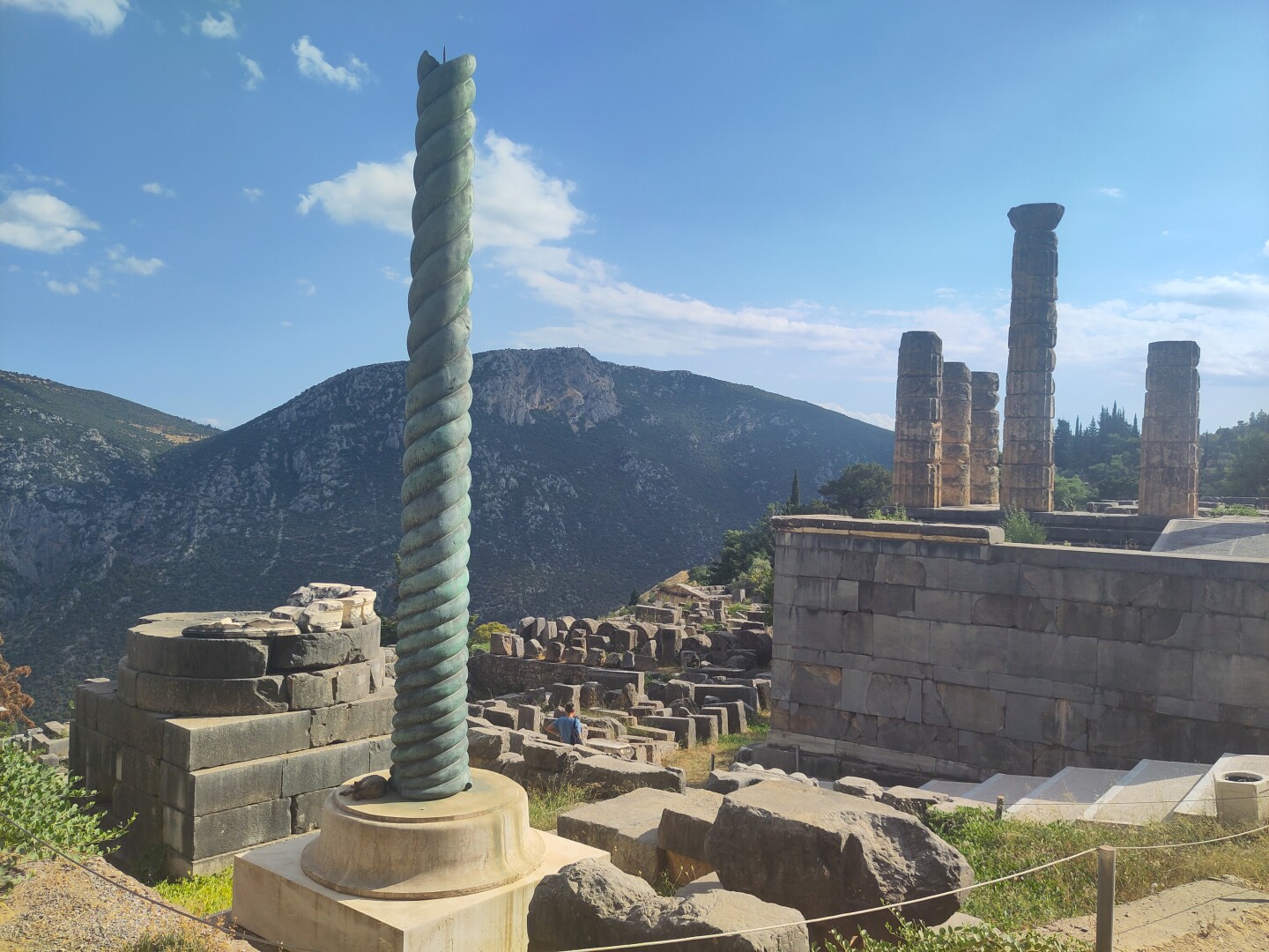 A tall spiraling bronze pillar takes up the foreground of the photo, with more of the Ancient Delphi ruins behind it with a mountain range in the back. To the right are the columns of the Temple of Apollo on the next level up.