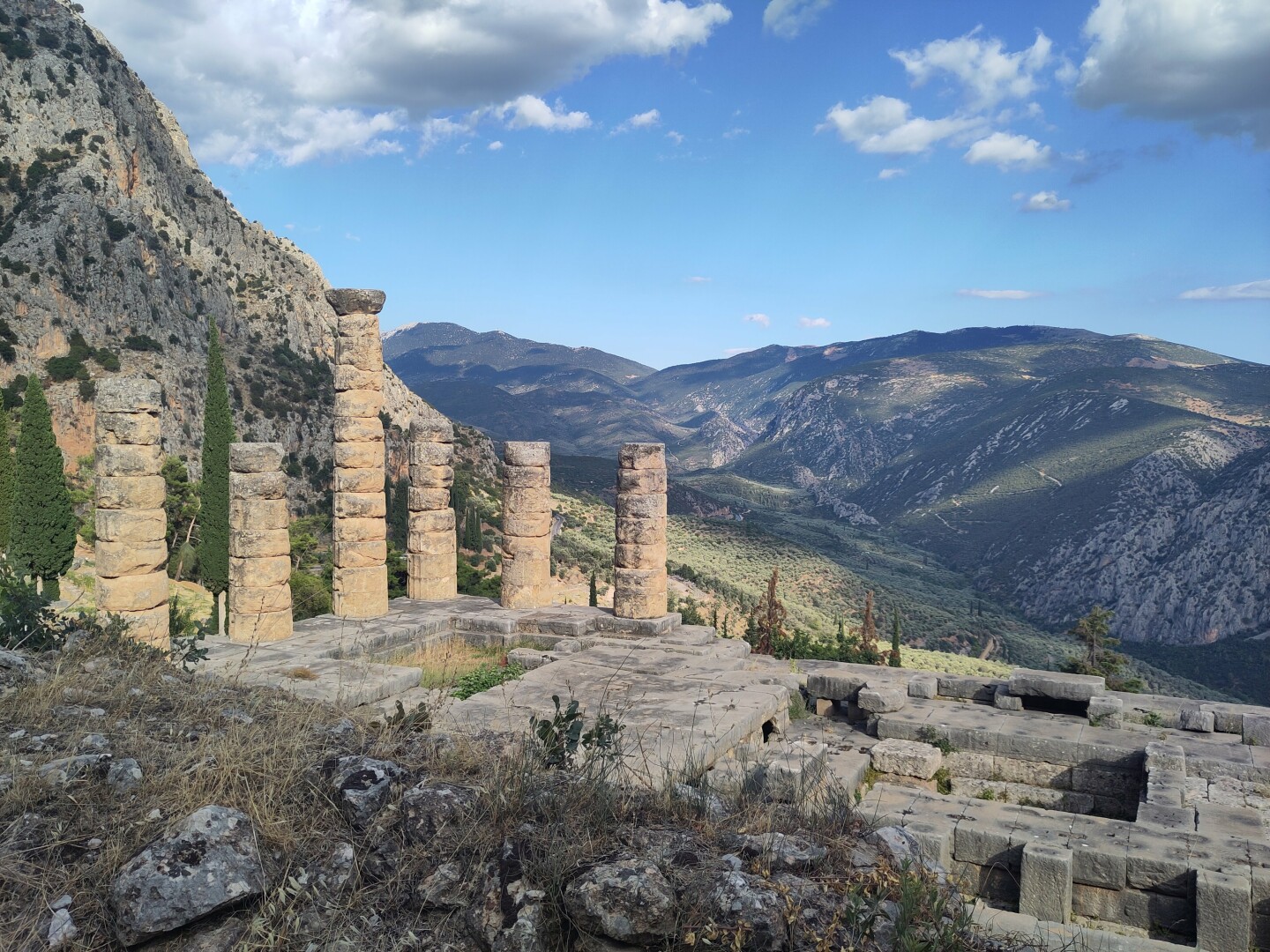 A broader landscape view of the Delphi ruins, centering a wider shot of the Temple of Apollo framed by the mountain range, with a bright blue sky above.