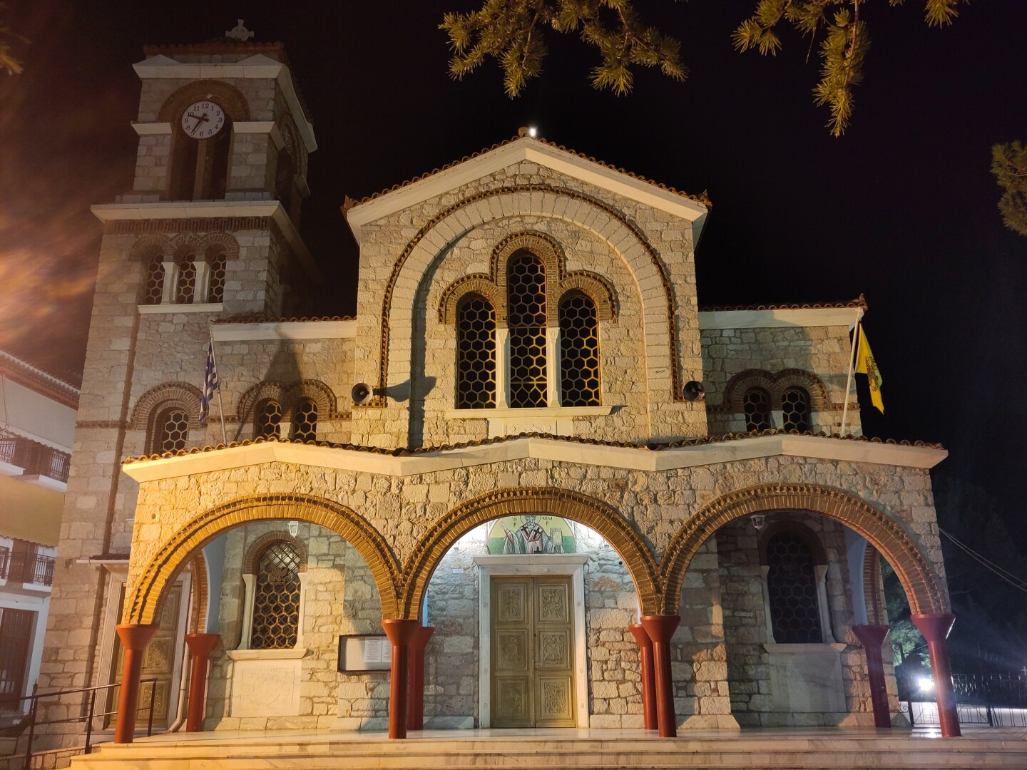 A night shot of the Church of St. Nicholas in Delphi, taken from the front. It has three arches making up the front gallery, a middle arch rising above that with a triple arched window inset, and a clock tower to the left. Some tree branches frame the right side of the building and an off-frame streetlight is causing a lens flare on the left.