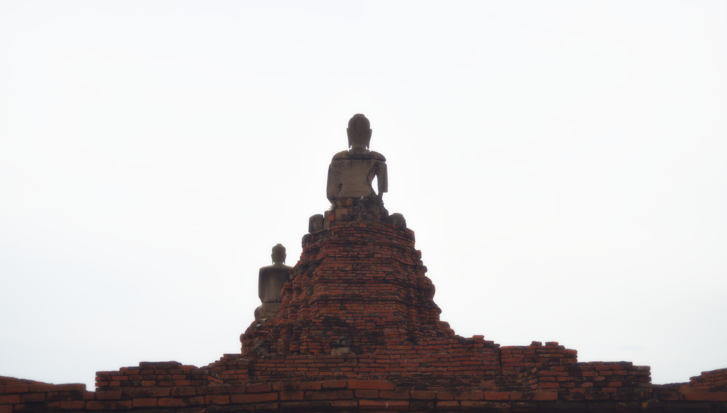Minimalist scene of old brick stones where one bigger and one smaller Buddha statues sit and meditate. They how the viewer their backs, the sky is overexposed, thus rendering totally white.