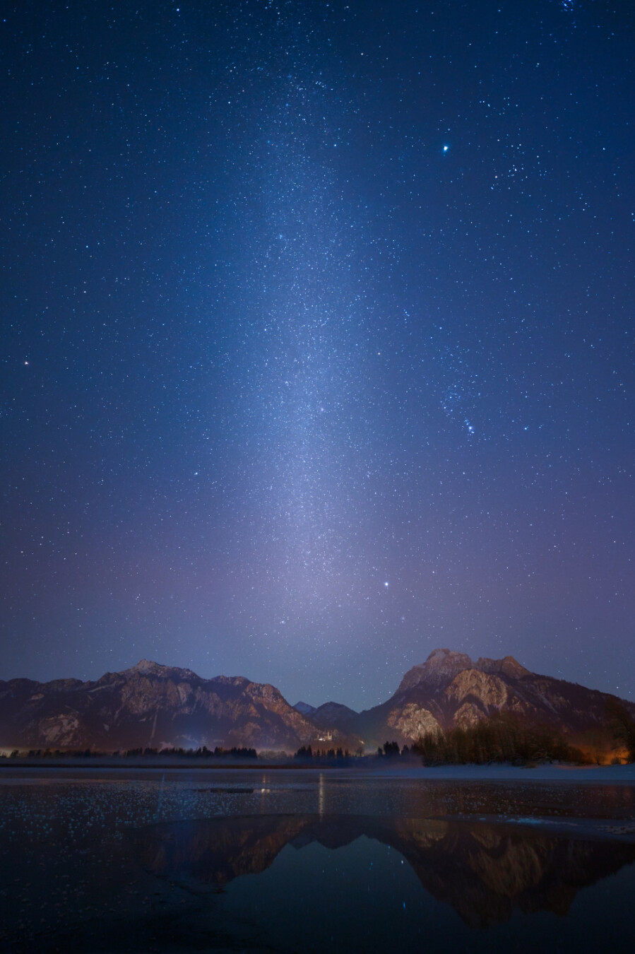 A vertical landscape shot at night showing the Forggensee, Bavaria with Neuschwanstein Castle in the middle. The upper part shows the vertical milky way, while the lake in the lower part is part frozen and has a reflection of the mountain range with the castle.