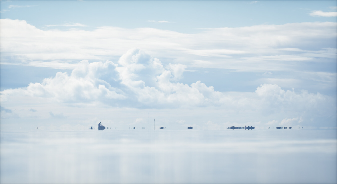 A colour photograph of the island of Utö and its lighthouse in the distance. The almost invisible horizon, dotted with some small islands and islets, lies in the lower third of the image. The calm sea reflects the soft white and blue tones of the summer sky. © Nika Potinkara