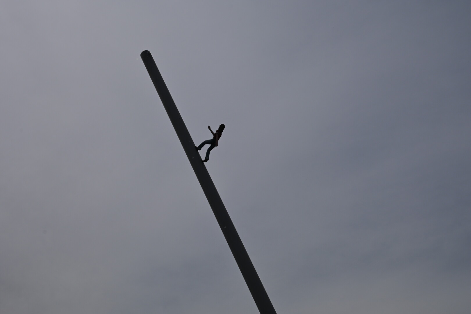 A silhouette figure is climbing a diagonally positioned pole against a cloudy sky. The figure appears to be defying gravity as they ascend the slanted structure.