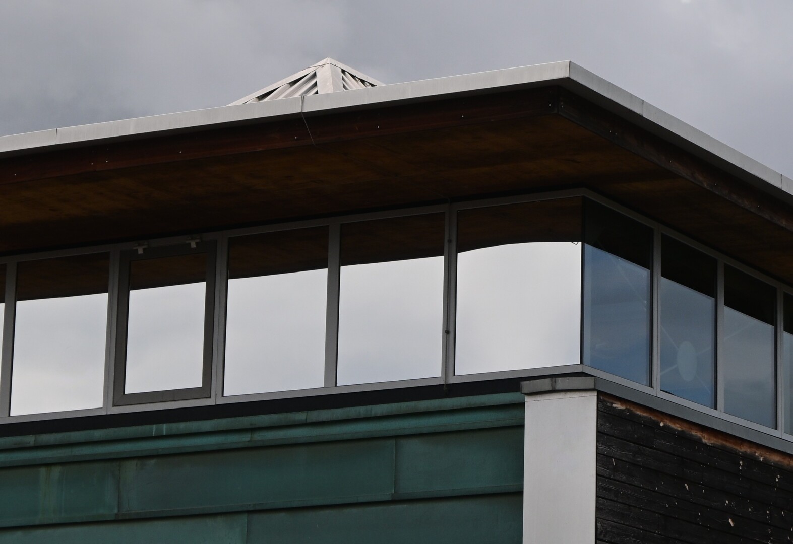 The image shows a modern building with large windows reflecting the cloudy sky. The building features wood and metal elements.