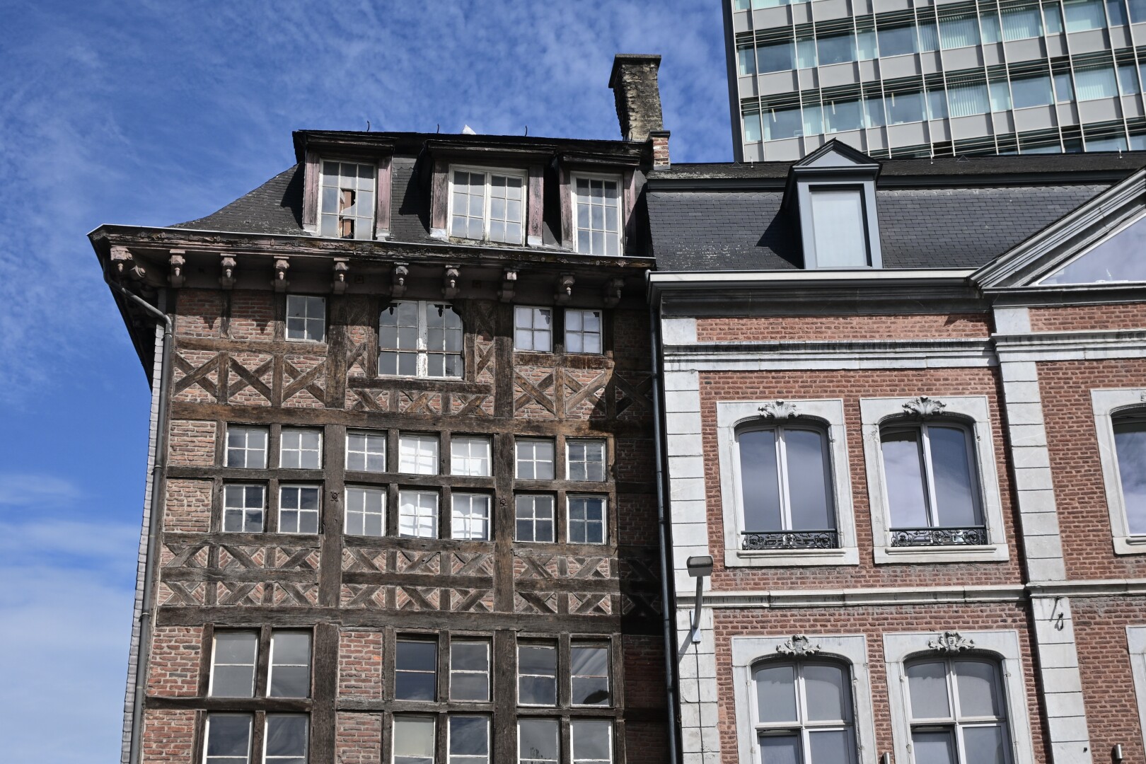 Two buildings with contrasting architectural styles stand side-by-side under a blue sky with wispy clouds. The building on the left is an older timber-framed structure with many windows, while the building on the right is a brick building with classical detailing. Peeking out from behind and above the older buildings is a modern, tall skyscraper. This building has a facade made of glass panels set within a grid of grey framing. Its sheer verticality and modern design contrast sharply with the older architecture below.