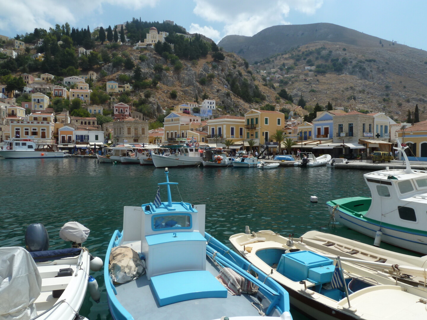 Pastel-coloured boats are moored in a harbour with pastel-coloured buildings climbing a hillside in the background. The sky is partly cloudy.