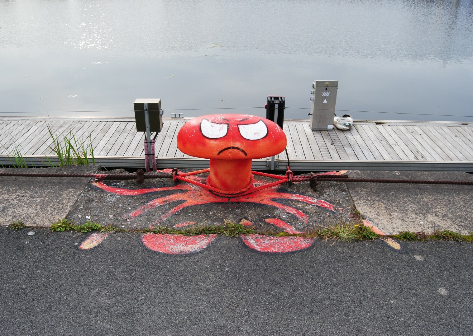A red, grumpy-faced bollard painted to resemble an octopus sits on a dock. Red paint spreads out from the base of the bollard, suggesting tentacles on the concrete ground.