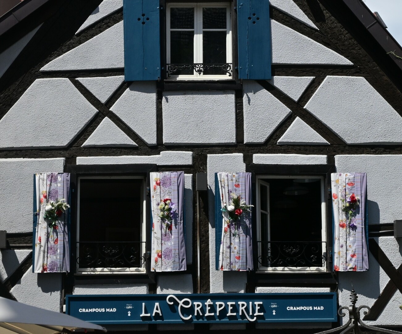 The image shows the facade of a creperie with traditional half-timbered architecture. There are two windows adorned with floral curtains and bouquets of flowers.