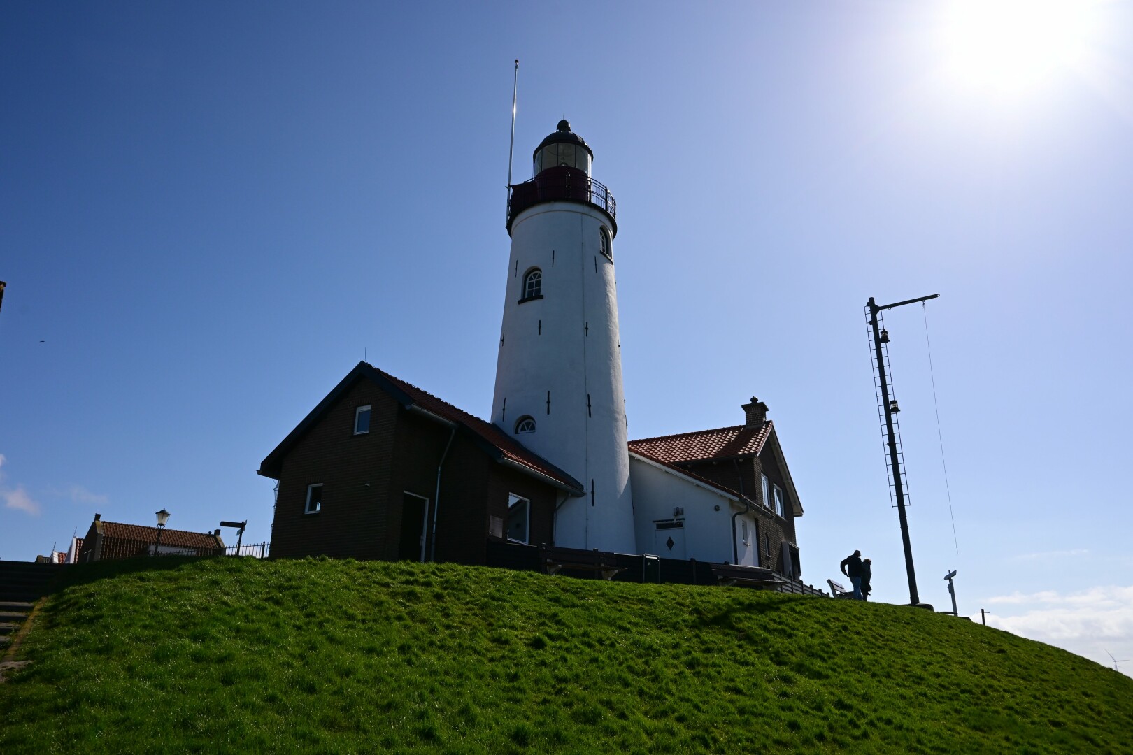 A white lighthouse stands on a grassy hill under a clear blue sky, with adjacent buildings and a tall pole with silhouetted figures. The sun is shining brightly, creating a strong backlight effect.