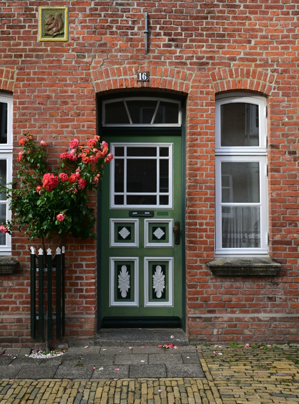 A red brick building features a green door with white decorative panels and a climbing rose bush. A window is visible on the right and a squirrel carving is above the door.