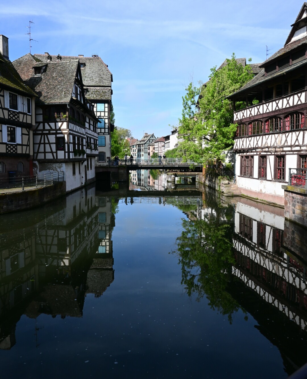 Traditional half-timbered buildings line a canal, their reflections mirrored in the calm water. Bridges span the canal, connecting the picturesque waterfront scenes.