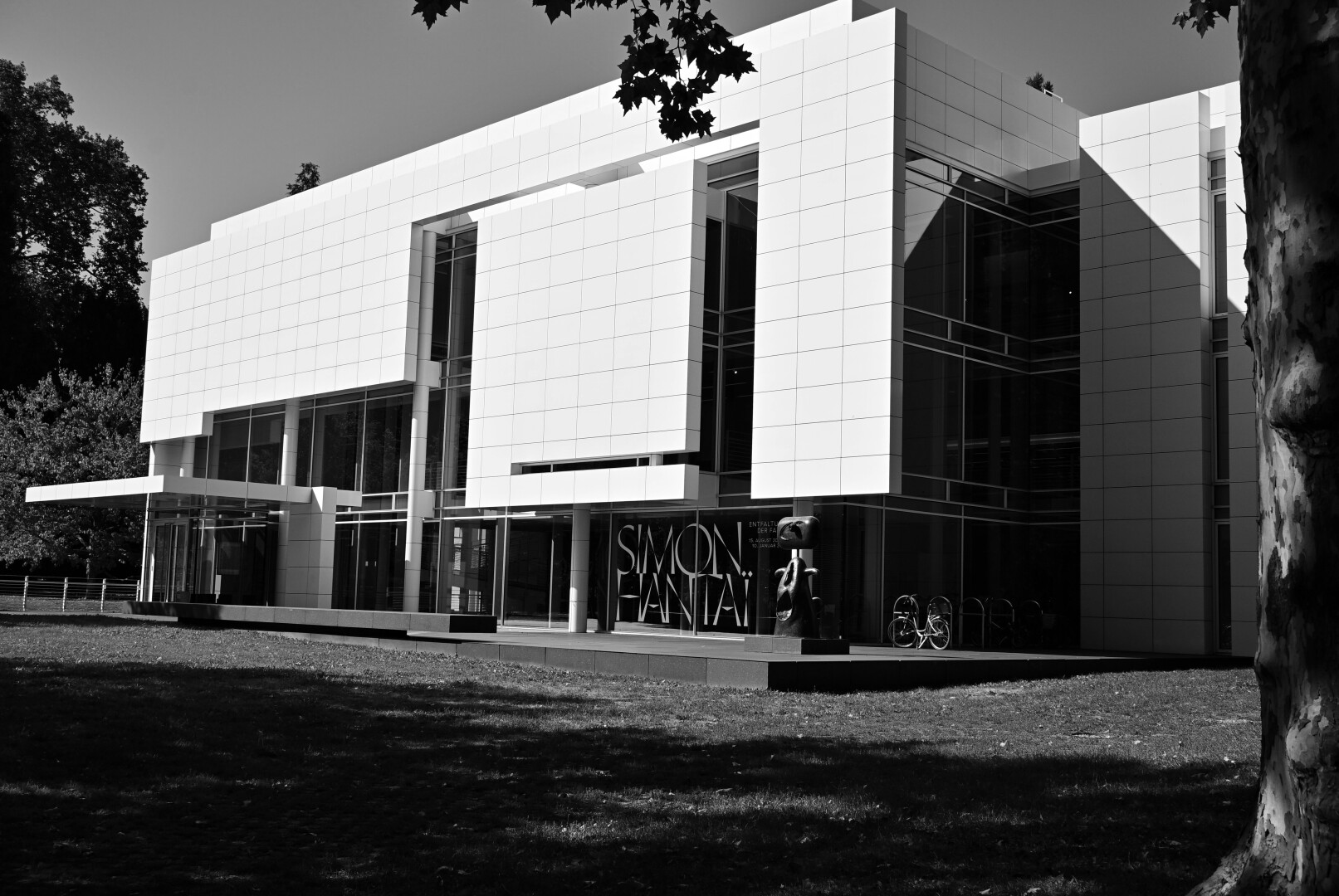 A modern, white building with large windows is shown in a black and white photograph. The building has "SIMON HANTAI" written on its facade, and a sculpture stands in front of it.