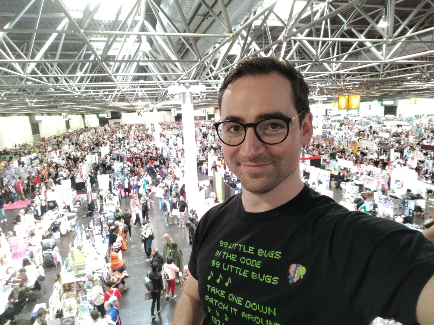 Selfie of myself in front of a big hall filled with thousands of people. I'm wearing a T-Shirt with a Developer Joke and a badge with a cute Cthulhu hugging a Blåhaj in front of a heart.