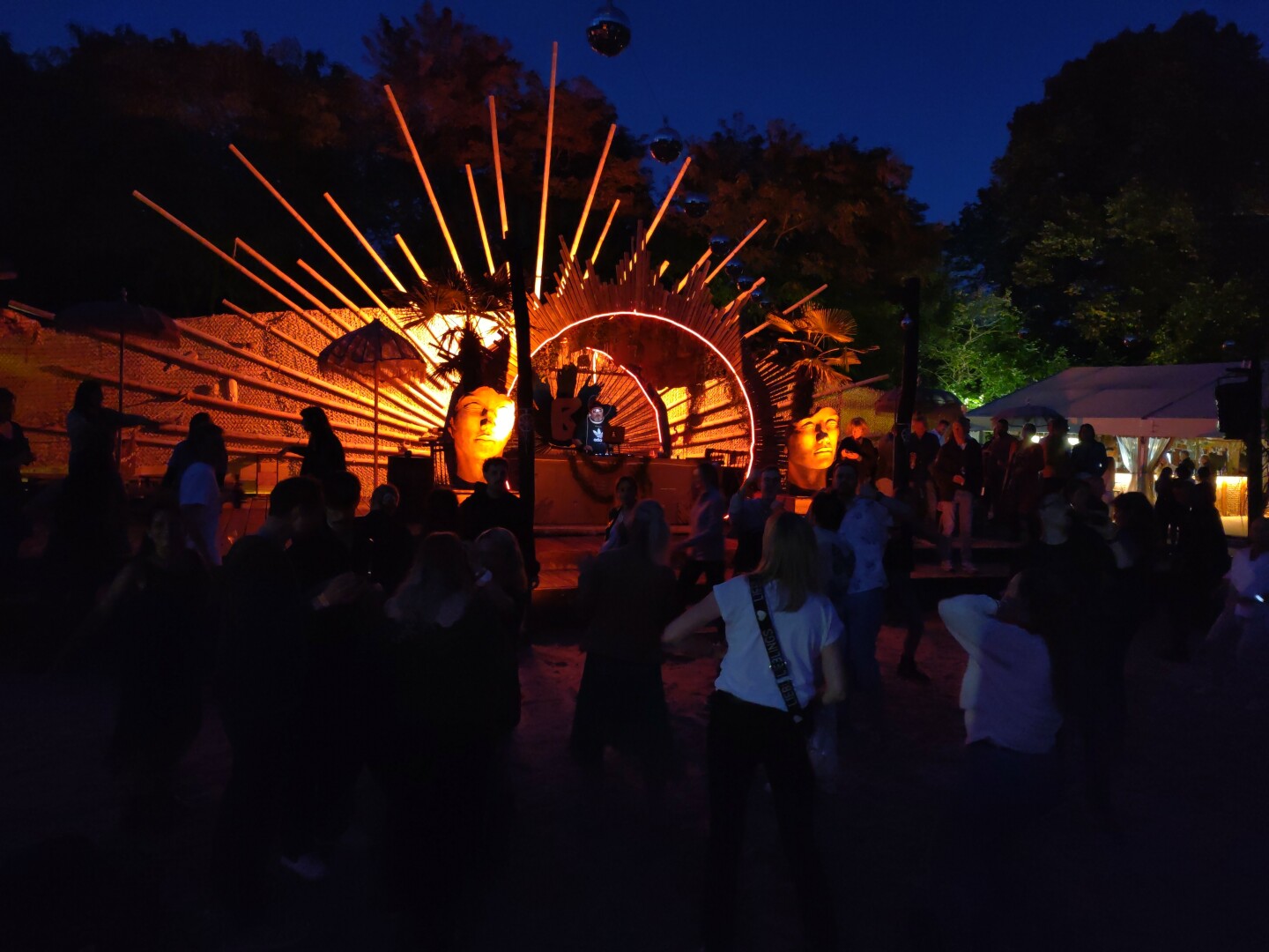 Photo of a beautifully lit DJ stage.
In front of the stage, there is a crowd dancing in the dark.